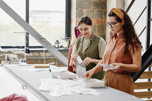 Contemporary young seamstresses in casualwear packing new shoulder pads for coats, jackets and dresses while standing by table in studio