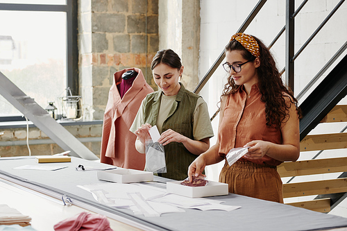 Two young seamstresses packing new shoulder pads for coats, jackets and dresses while standing by table in studio or workshop