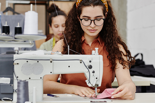 Young serious seamstress sitting by electric sewing machine while making shoulder pads for new dress, coat or other clothing item