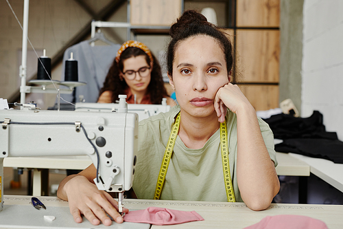 Young tired or bored seamstress with measuring tape on her neck looking at you while sitting by electric sewing machine in workshop