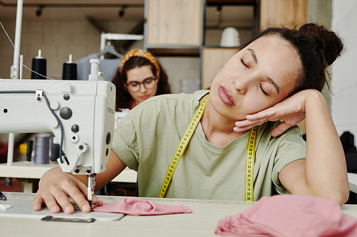 Young tired seamstress with measuring tape on her neck sitting by electric sewing machine with her eyes closed and having short break