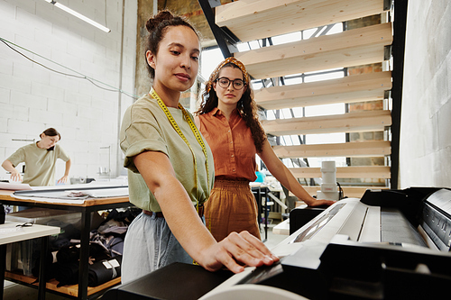 Two young tailors standing by large industrial printer under staircase in studio or workshop and printing sketches of new clothing items