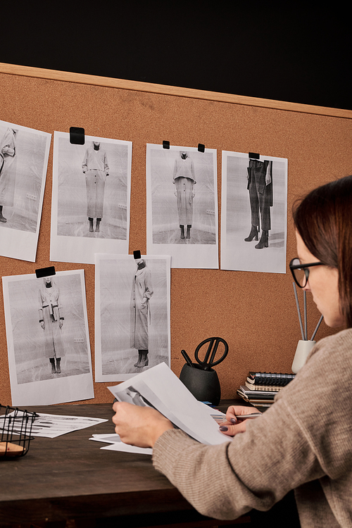 Young brunette female fashion designer working with photos while sitting by table in front of carton board with group of trend examples