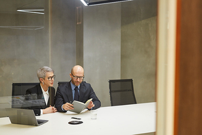 Wide angle portrait of two successful mature business people reading notes while sitting at table in minimal office interior behind glass wall, copy space