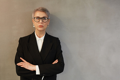 Waist up portrait of successful mature businesswoman standing with arms crossed and looking at camera while posing against minimal gray wall and wearing official suit, copy space