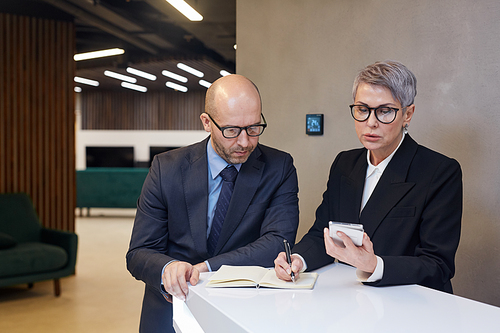 Portrait of two mature business people writing out data from smartphone at stand in minimal office interior, copy space