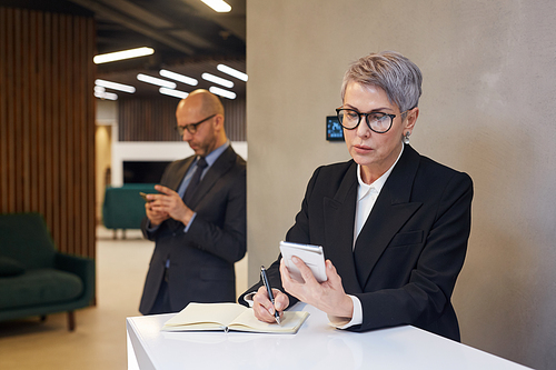 Waist up portrait of mature businesswoman writing out data from smartphone at stand in minimal office interior, copy space