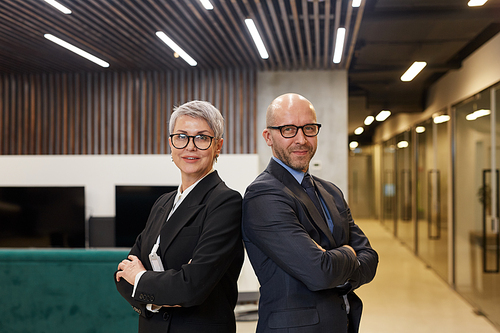 Waist up portrait of two successful mature business people standing back to back with arms crossed and looking at camera while posing in modern office interior, copy space