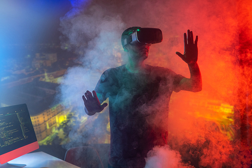 Contemporary young male it-manager with vr headset touching virtual display while standing in smoke on red lit background