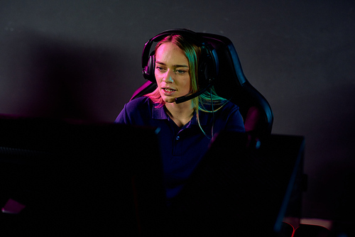 Young serious blond female e-sport gamer in headset looking at computer screen while sitting in dark club in front of monitor
