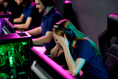 Tired or failed girl in headset touching her head while bending over desk in front of computer monitor on background of video gamers