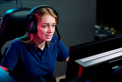 Young female e-sport addict in headset looking at computer screen during cyber competition while sitting in modern game club