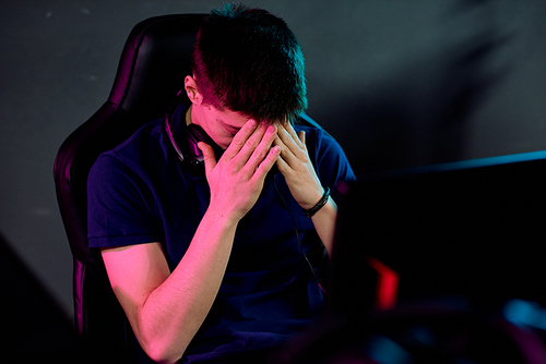 Worried young man with headphones around neck covering his face by hands during cyber sport game while sitting in front of monitor