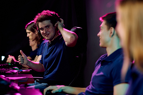 Young successful e-sport gamer showing thumb up and looking at one of friends with smile while playing cyber game in front of monitor