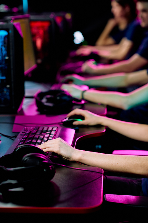 Row of hands of young contemporary e-sport clubbers sitting by desks in front of computer monitors and playing cyber games