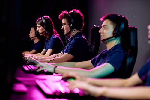 Row of guys in casualwear and headsets sitting in black leather armchairs in front of computer screens and playing e-sport games in club