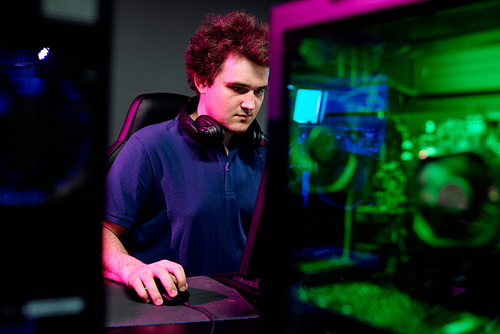 Serious guy with headphones around neck looking at computer screen during e-sport cyber game while playing in contemporary club