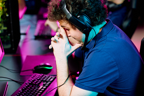 Tense or worried young man in headset touching face while slightly bending over keyboard in front of computer monitor