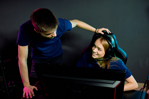 Young e-sports participant standing at armchair of female gamer and talking to her about videogame progress