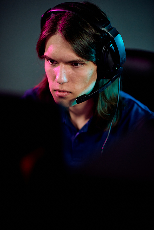 Focused guy with long hair using headset with microphone for communication with network gamers while playing computer game