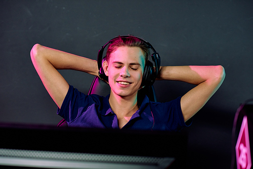 Young smiling relaxed male video gamer in headphones enjoying break between e-sport games while keeping his hands behind head