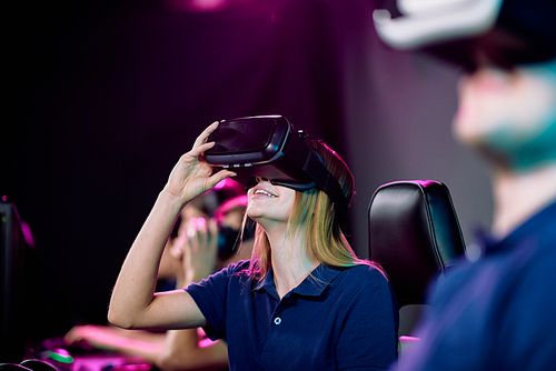 Happy young female video gamer in vr headset sitting among her friends in cybersports gaming club and enjoying network game