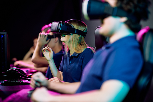 Teenage cybersports gamer in virtual reality goggle watching and taking part in network video game with her friends sitting near by