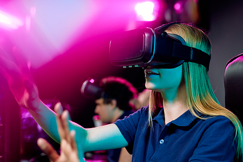 Blond teenage girl in vr goggle touching virtual display while taking part in cybersports video game on background of her friends