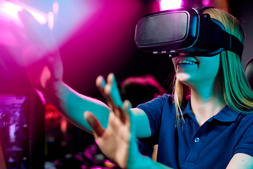 Positive girl using virtual reality simulator gesturing hands to bright purple light while playing video game in 3D