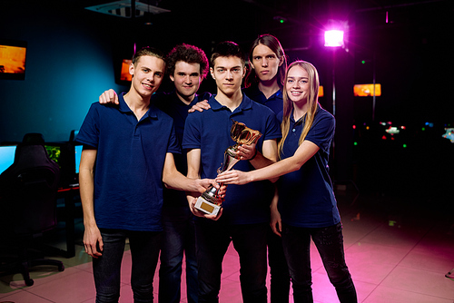 Five young cheerful and successful e-sports competition winners holding golden award statue while standing in front of camera in club