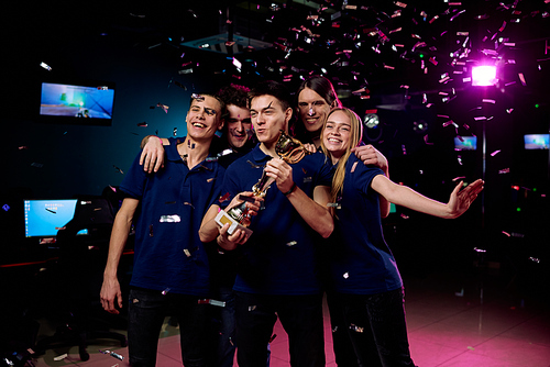 Group of five teenagers celebrating their victory in cybersports competition while standing in confetti fall and showing golden cup