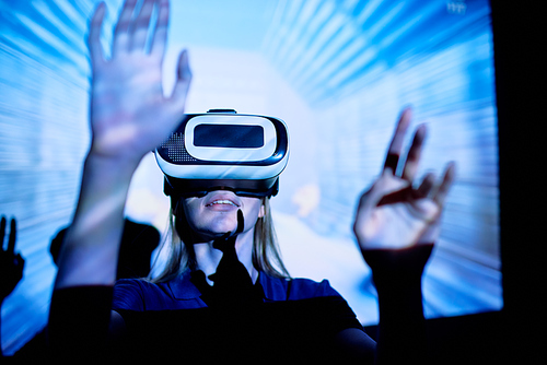 Young woman standing in blue light and playing virtual game in VR goggles, virtual 3D picture showing on projection screen