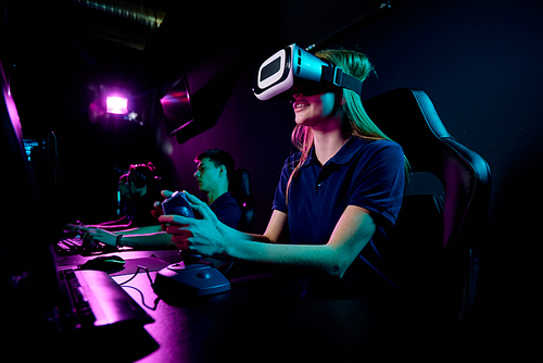 Contemporary eenage girl in vr headset using joystick while sitting in front of computer monitor and playing new video game in club