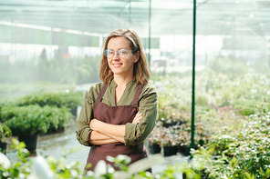 Happy mature female gardener in eyeglasses and workwear crossing arms by chest while standing by large flowerbed with green plants