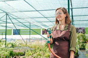 Happy mature brunette female gardener in eyeglasses and brown apron watering plants in hothouse while standing against large flowerbed