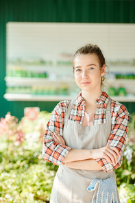 Young serious female farmer or greenhouse worker in workwear standing in front of camera and looking at you against green plants