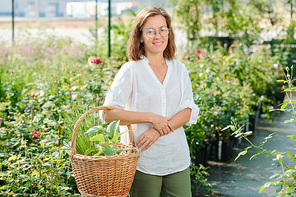 Cheerful brunette female worker of hothouse or gardener in casualwear and eyeglasses holding basket with green plants against flowerbeds
