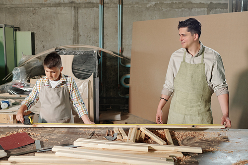 Concentrated teenage boy in apron standing at desk with wooden pieces and assisting father to measure plank
