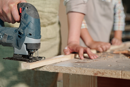 Close-up of unrecognizable carpenter holding plank and using jigsaw while cutting it in workshop