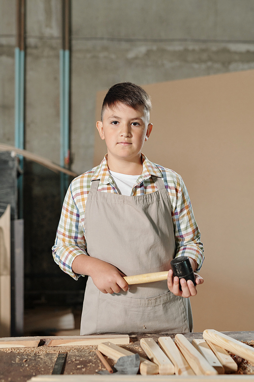 Portrait of serious teenage boy in apron standing with hammer at desk with wooden planks in carpentry shop
