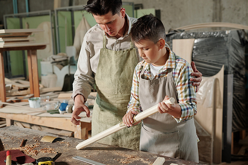 Teenage boy in apron making plank smooth with sandpaper under control of father in carpentry workshop
