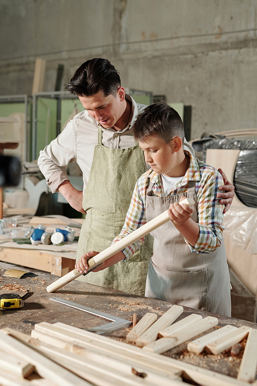Skilled carpenter in apron supporting son while he learning to polish wood with sandpaper in workshop