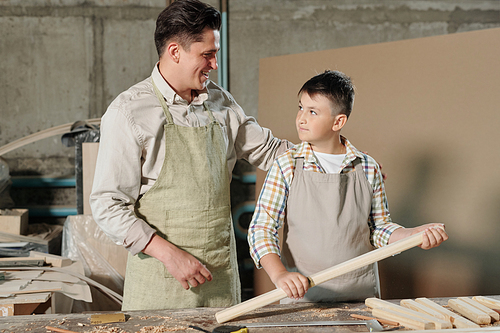 Satisfied middle-aged carpenter in apron being proud of teenage son praising him for good job in workshop