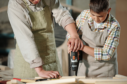 Close-up of skilled carpenter in apron pushing hands of son while helping him to drill wooden surface in workshop