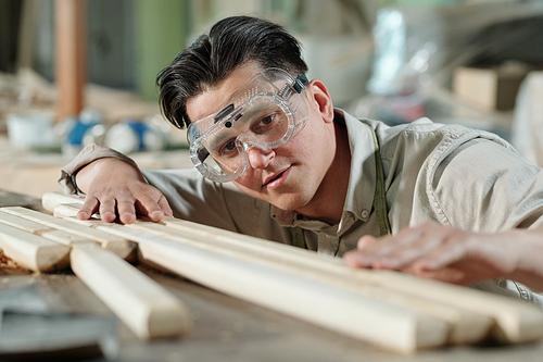 Concentrated middle-aged male carpenter in safety goggles examining surface of wooden planks in workshop