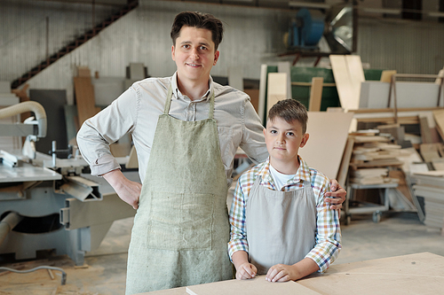 Portrait of smiling middle-aged carpenter in apron embracing teenage son in furniture workshop