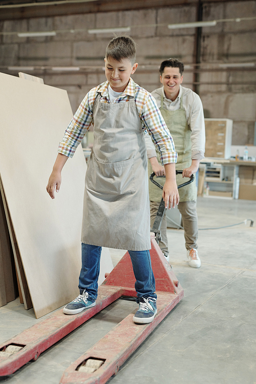 Cheerful middle-aged father pushing trolley jack with son while having fun in furniture workshop