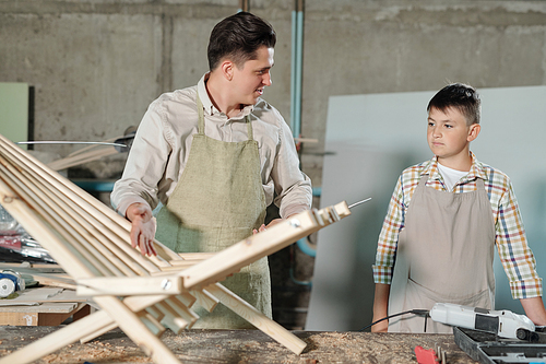 Middle-aged father talking to teenage son while they making wooden chaise lounge in furniture workshop