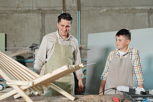 Smiling middle-aged father and son in aprons looking at wooden chair in carpentry workshop