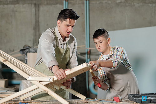 Skilled teenage boy in apron using hammer while working on wooden furniture with father in carpentry shop